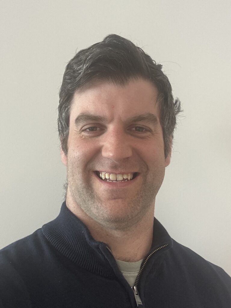 Close-up of a man smiling at the camera, wearing a dark zip-up jacket against a light background.