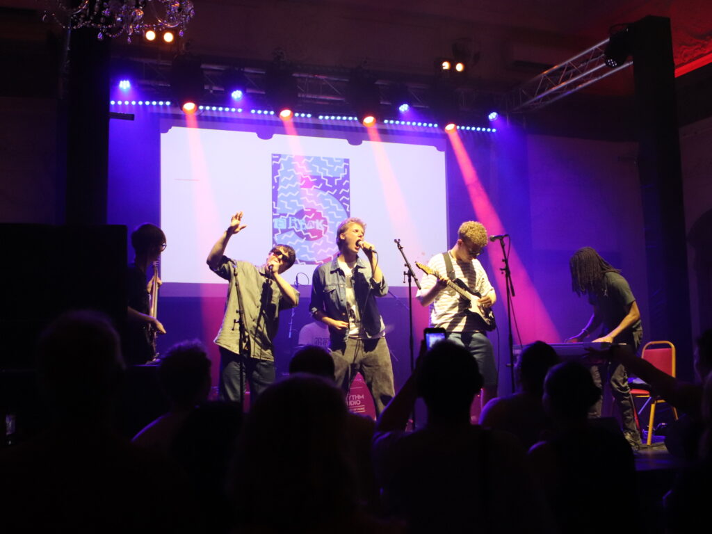 Band performing on a lit stage with colorful purple and red lights, four musicians singing and playing guitars, audience silhouetted in front.